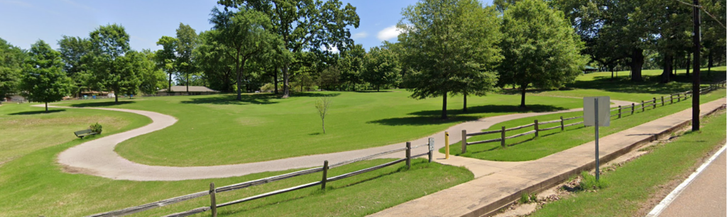 Photo of a park on a sunny day with a blue sky and a green lawn, scattered trees, and a winding concrete walkway behind a split-rail wooden fence. 