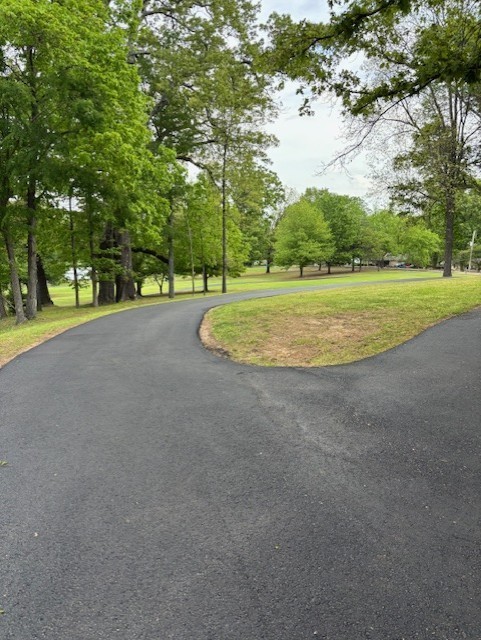 Freshly paved black walking trail running through a park of green grass and trees. The trail splits and heads down a slight hill or up to the right. 