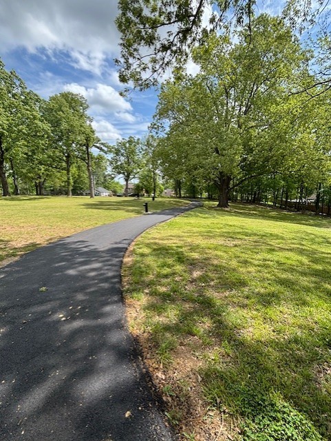 Freshly paved black walking trail running through a park of green grass and trees. A blue sky with white clouds is above