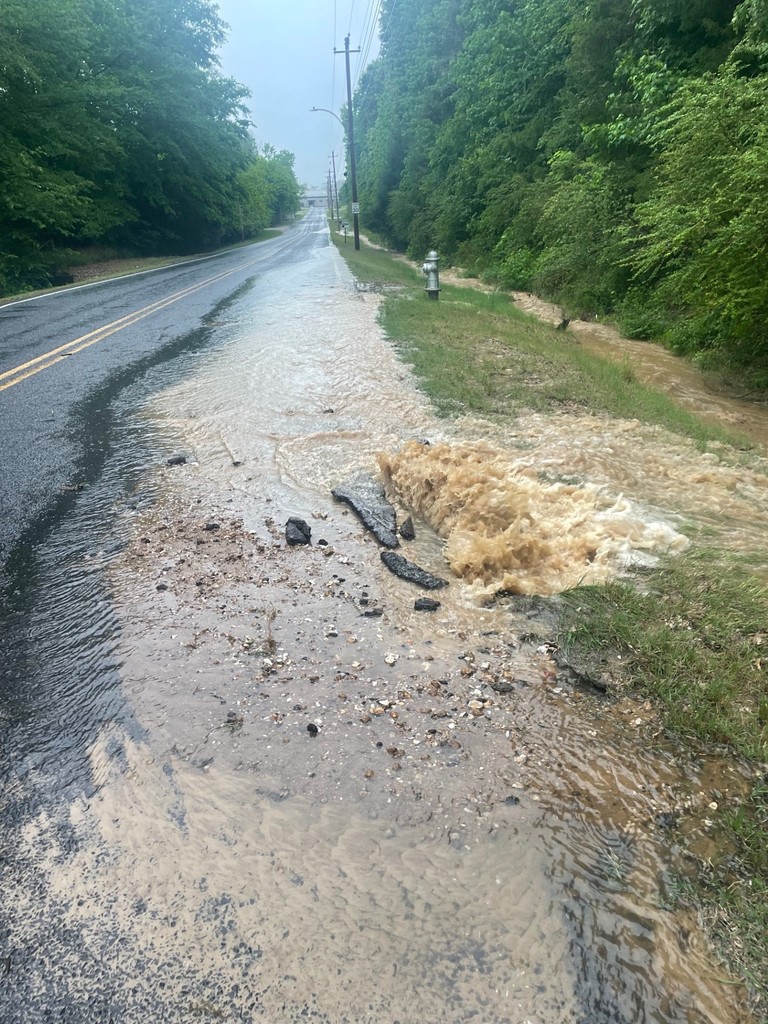 Picture of brown water bubbling up through a hole in the side of a 2-lane road on a rainy day. Water is running into a ditch, which is full of muddy water and flowing off into the distance. 