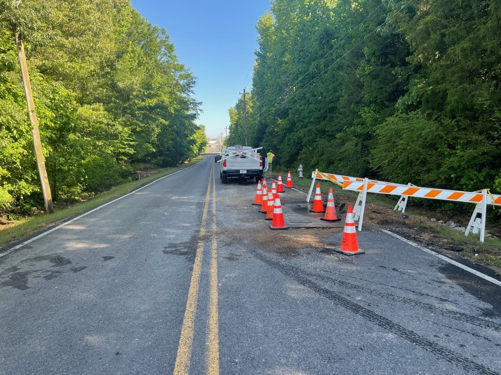 Picture of 2-lane road lined with trees. The road has a metal plate on the right lane surrounded by orange cones and two orange and white barriers are on the shoulder around a dirt area of recent repairs. 