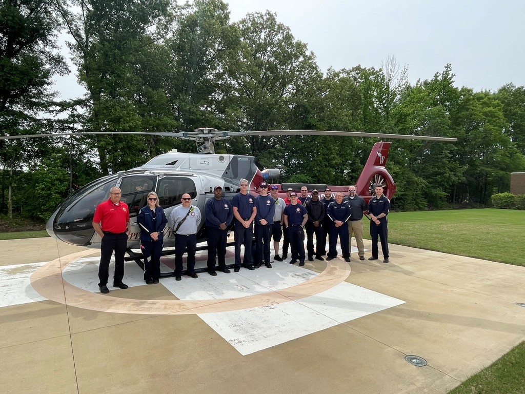 Picture of a line of firefighters posing in front of a hospital helicopter on a landing pad with a line of trees in the background. 