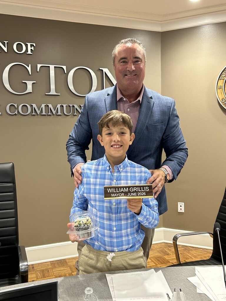 Mayor Wissman and student William Grillis posing at the front of the Arlington Board Room. William is holding a cupcake and his name plate as Mayor for the Day