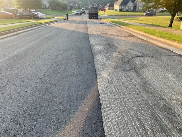 photo of a 2-lane street with one lane freshly paved and the other milled. in background is a dump truck and construction worker in reflective vest.