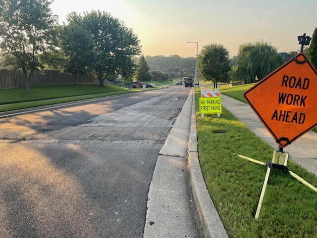 Photo of a 2-lane street with one side milled. In foreground is an orange Road Work ahead sign, and a yellow "no parking, street paving" sign.