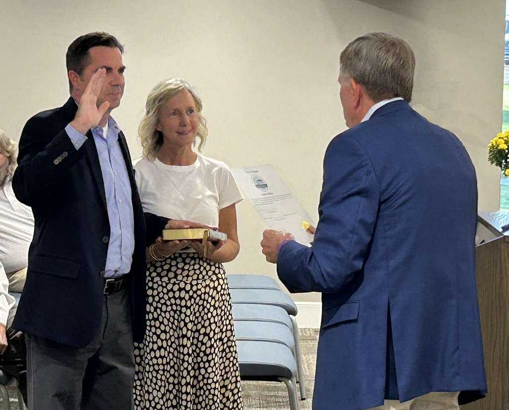 Alderman Biggs, in a dark jacket and blue shirt, is taking the oath of office with one hand on a bible held by his wife and the other raised. Senator Rose in a blue jacket is administering the oath.