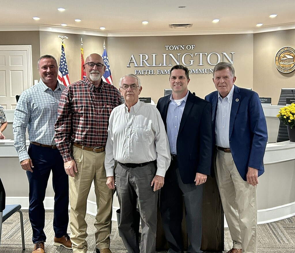 Five men in suits posed in front of the Town of Arlington Board Room, with US and TN flags, a round Town seal, and signage for the Town in the background.