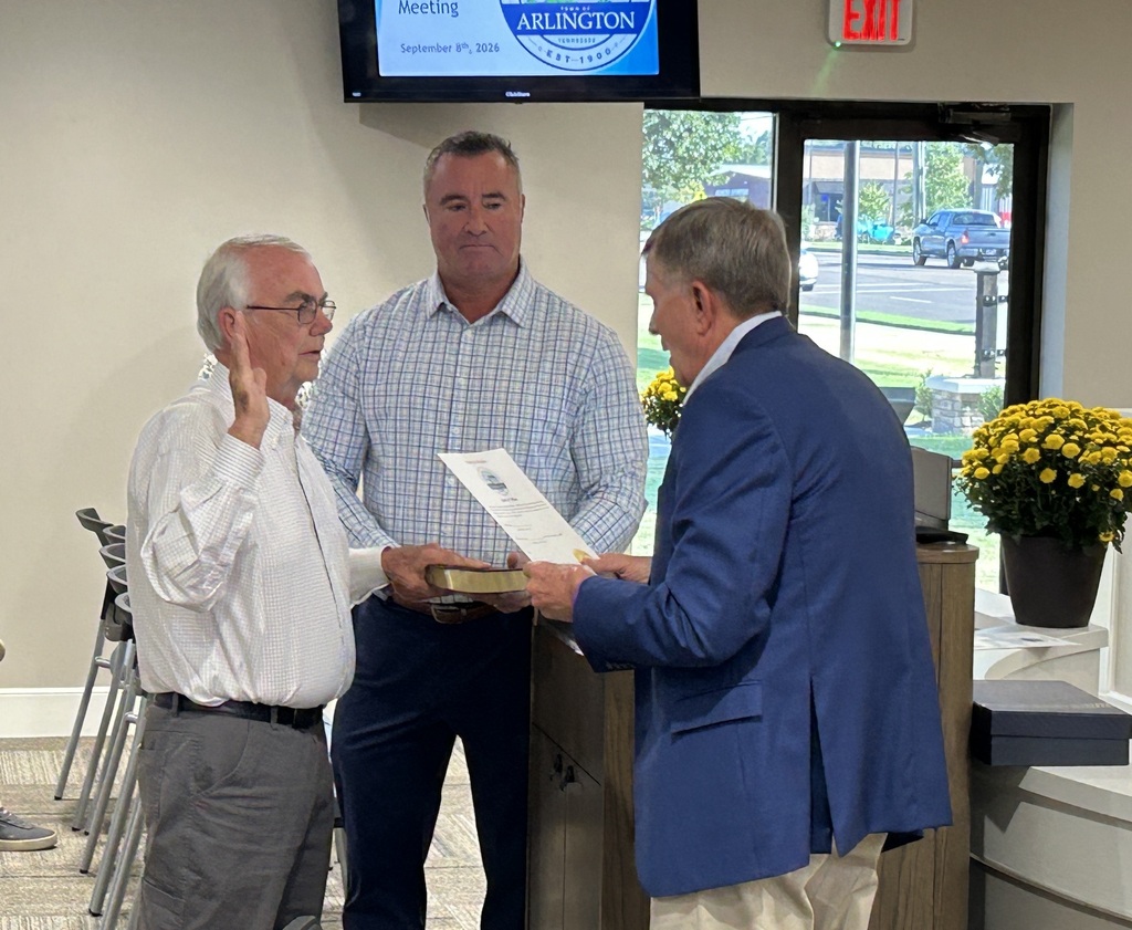Alderman Harry McKee taking an oath of office, with one hand on a bible and the other raised. Senator Rose in a blue suit jacket is reading the oath, while Mayor Wissman in a button down shirt is holding the bible.
