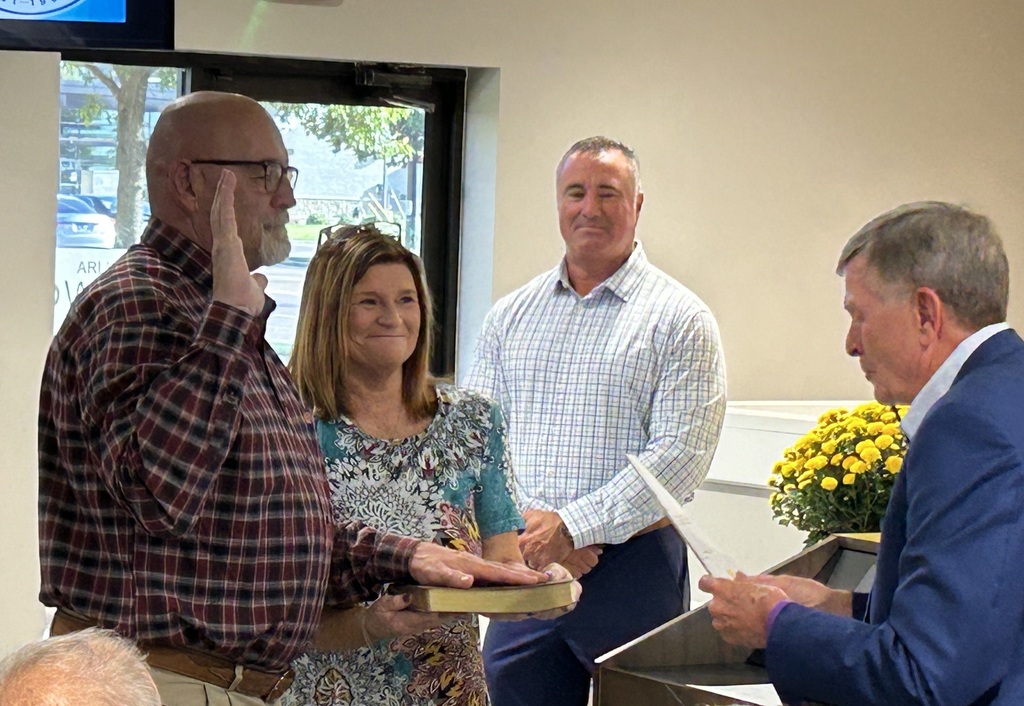 Alderman Brooks in a red and blue button down shirt is taking the oath of office, with one hand on a bible held by his wife and the other raised. Senator Rose is reading the oath, while Mayor Wissman in a blue and white button down is in the background