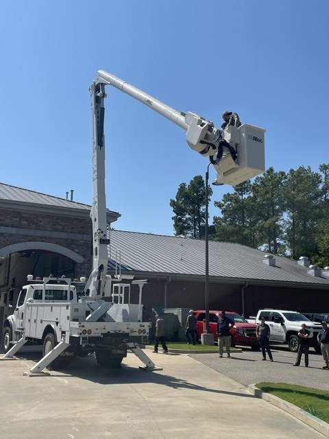 Photo of a new white bucket truck parked in front of a fire station with the stabilizers out and two people in a bucket raised up in the air. A group of others are standing below and watching.