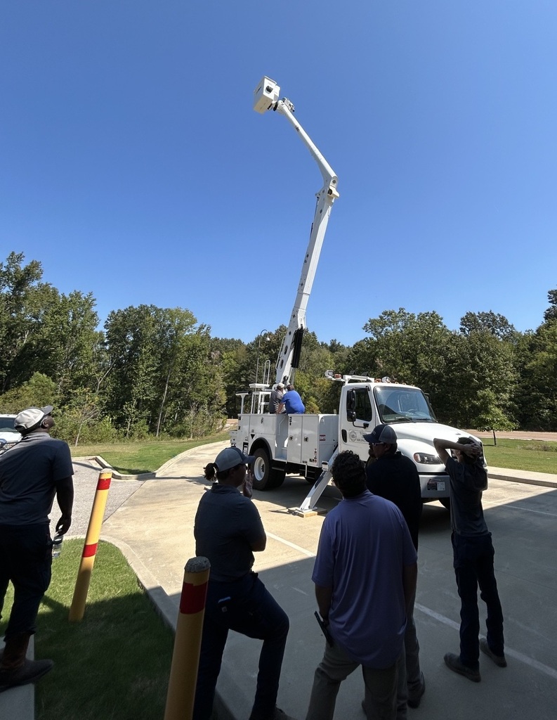 Photo of a white bucket truck with arm fully extended toward the bright blue sky, while a group of employees look on from below