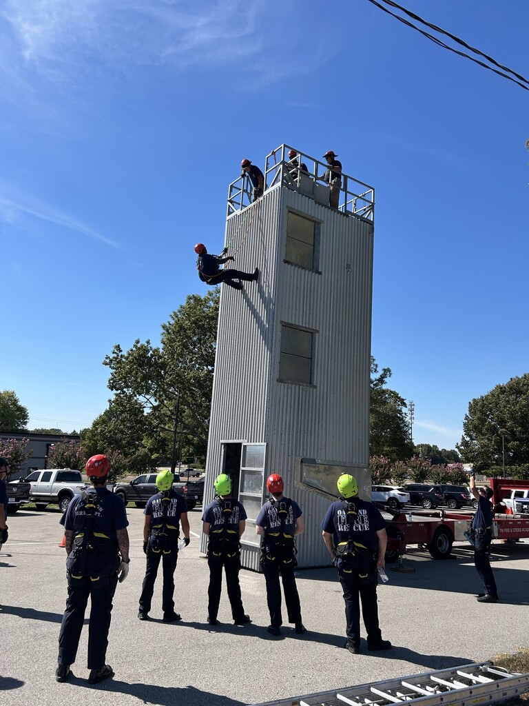 Photo of a firefighter in blue decending off the top of a 3-story metal, mobile practice tower, with bright blue skies in the background and several firefighters waching from below