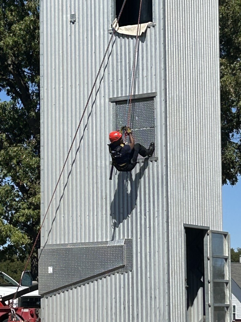 Photo of a firefighter in blue with a red helmet hanging from ropes as he climbs up the side of a metal practice tower