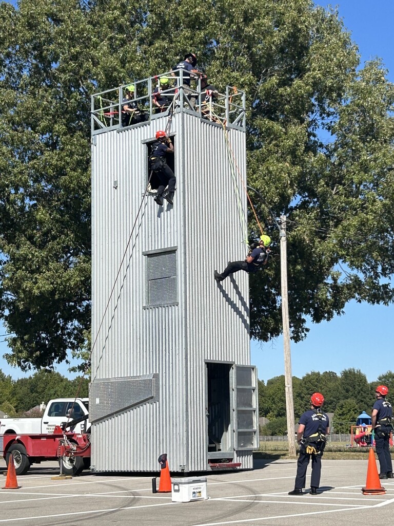 Photo of a 3-story mobile, metal practice tower with firefighters on top, two on the sides hanging from ropes, and several on the ground watching them ascend and descend.