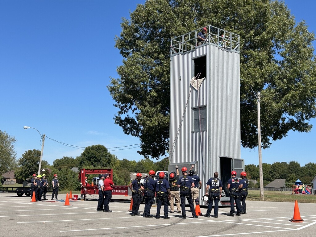 Photo of a group of firefighters in harnesses and red helmets looking up at a 3-story metal training tower in a parking lot, with a large tree and blue sky in the background