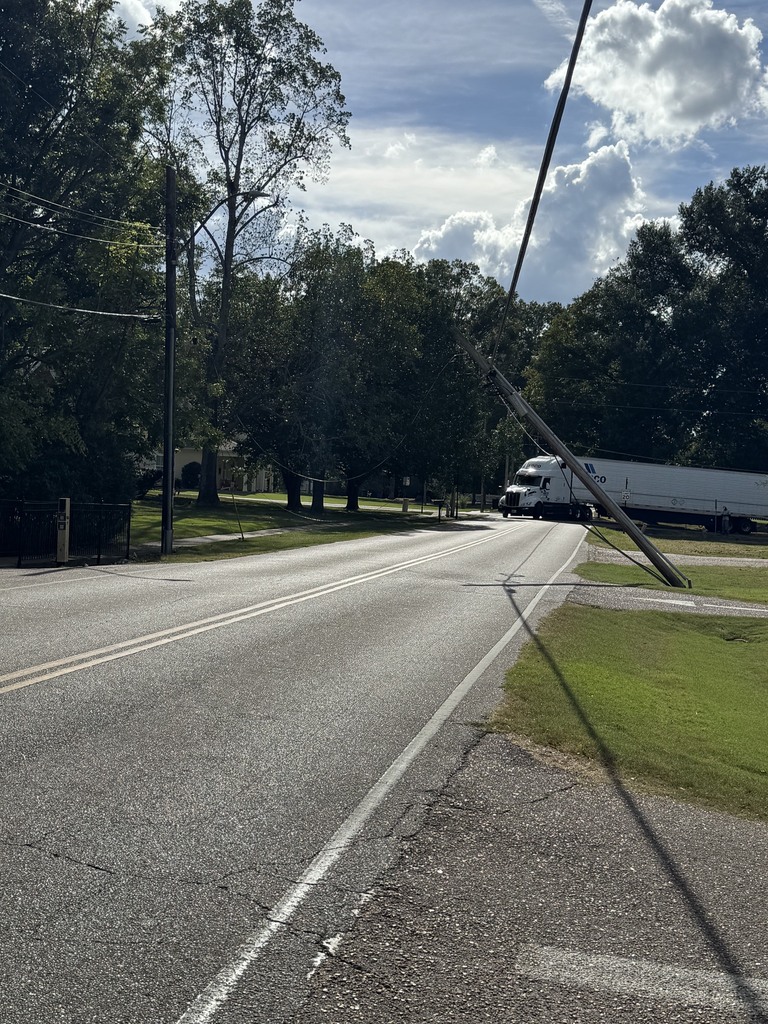 Photo of a 2-lane road with a large white semi-truck in the background that is clearly caught on overhead power lines. A pole in the foreground is pulled and leaning over the roadway