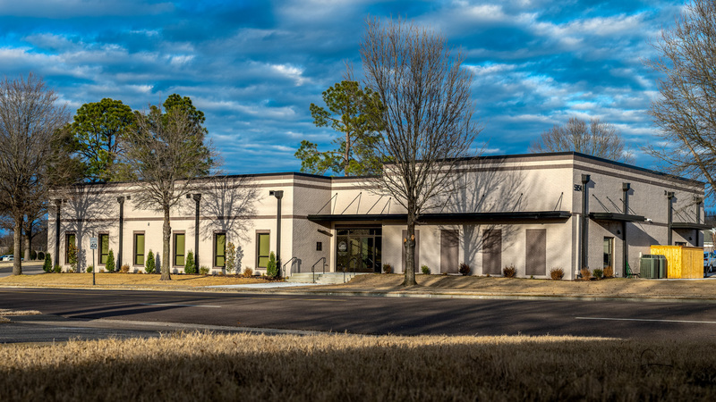 Photo of single-story beige brick building with dark bronze trim and awnings. Includes a line of windows along the front and glass double doors in the center. Blue sky above and trees around the perimeter.
