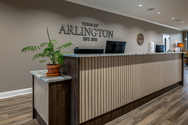 Photo of a front reception counter with beige wall behind it. On the wall is raised lettering: Town of Arlington, est 1900.