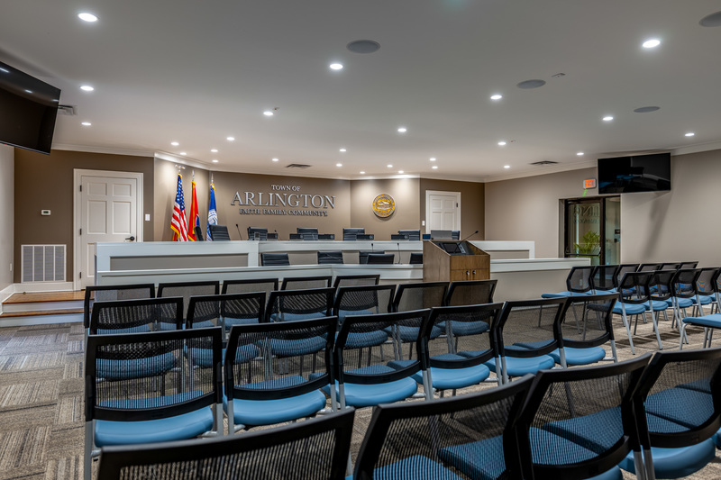 Photo of a board room with lines of blue and black chairs, and a raised dais with light brown wall behind it with raised letters that say Town of Arlington, flanked on either side by 3 flags on left and a round town seal on right