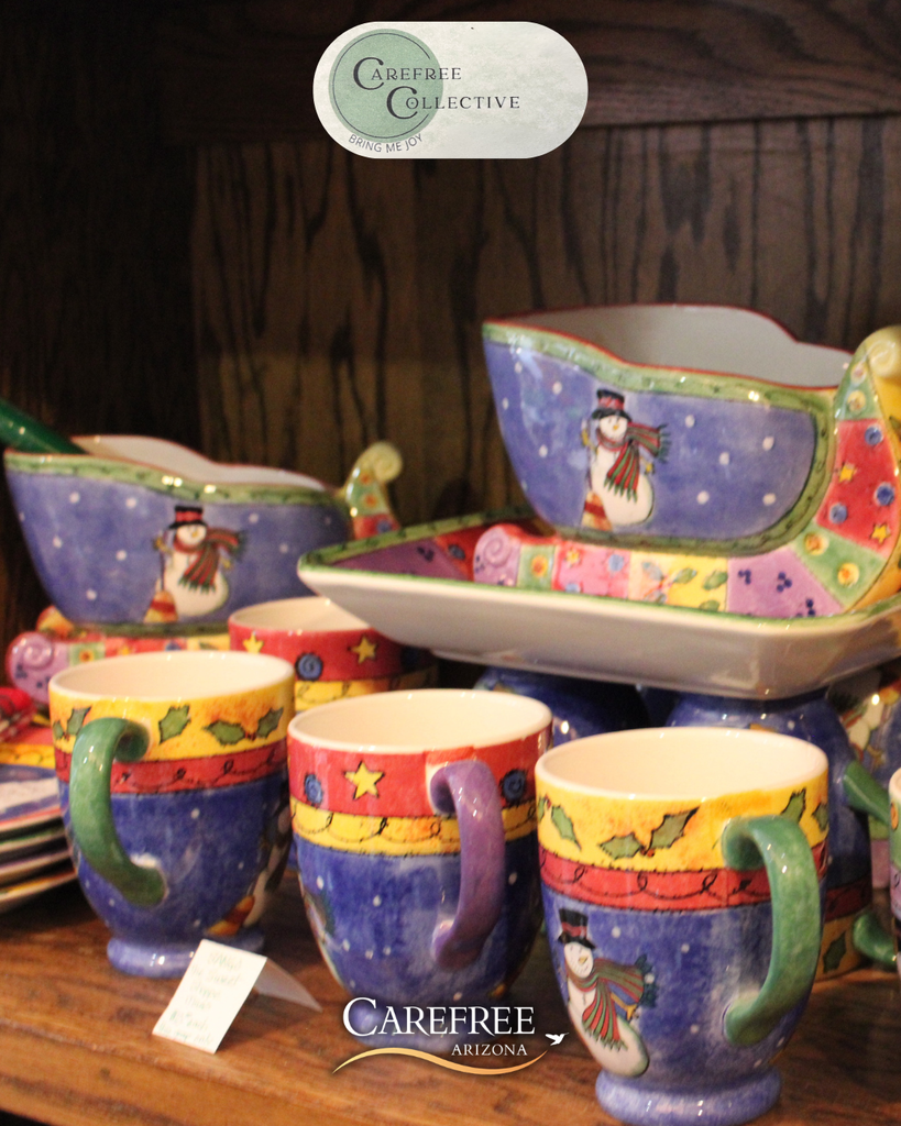 Colorful ceramic mugs and bowls decorated with snowmen and holiday motifs on a wooden shelf.