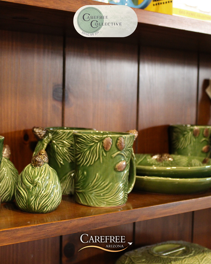 Green ceramic mugs and plates with pinecone and pine needle designs displayed on a wooden shelf.