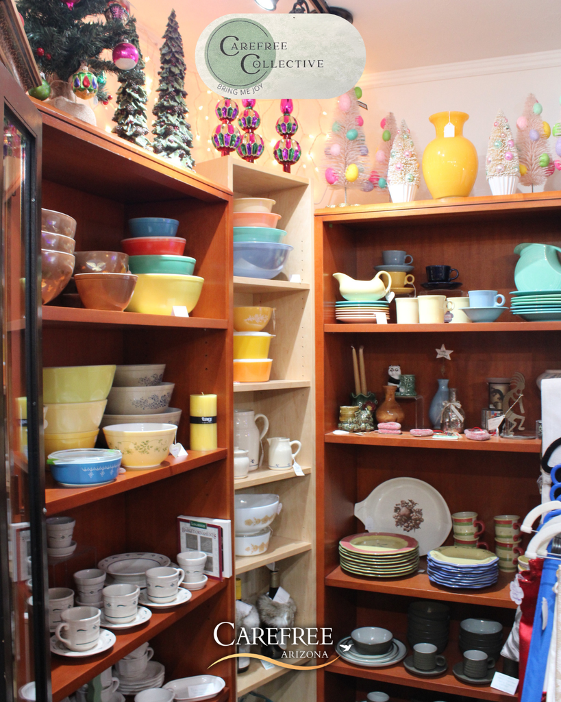 Shelves filled with colorful vintage Pyrex bowls, plates, and cups in a cozy shop corner decorated with pastel Christmas trees and ornaments.