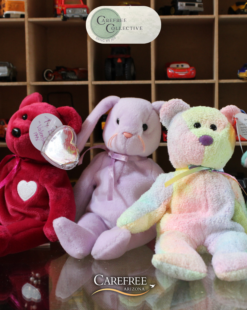 Three Beanie Babies in red, lavender, and pastel rainbow colors sit on a reflective surface with toy cars displayed in cubby shelves behind them.