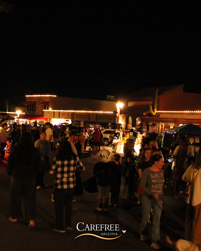 Crowds of people walk and gather at a nighttime outdoor event in Carefree, Arizona, with buildings lit by string lights in the background.
