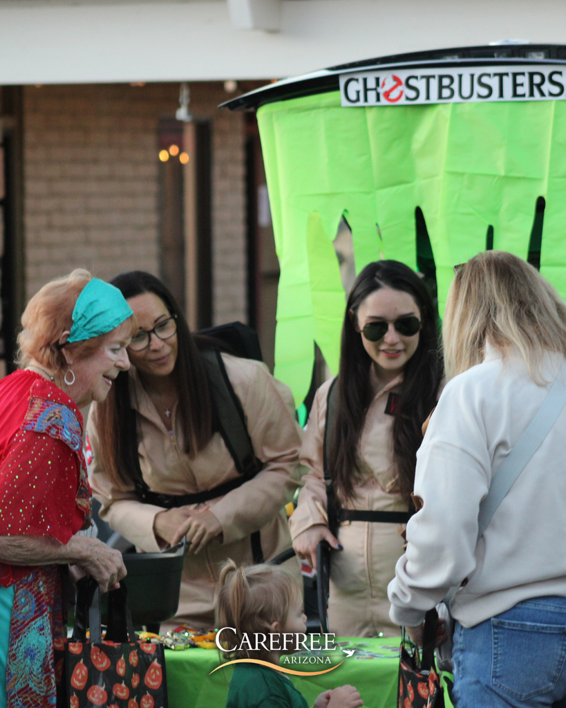 Two women dressed as Ghostbusters interact with a group at a festive Halloween-themed table outdoors.
