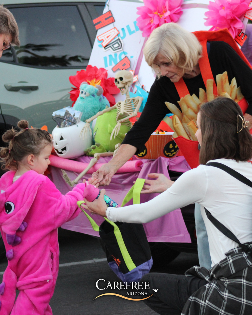 Child in pink dinosaur costume receiving candy from an older woman dressed as french fries during a Halloween event.