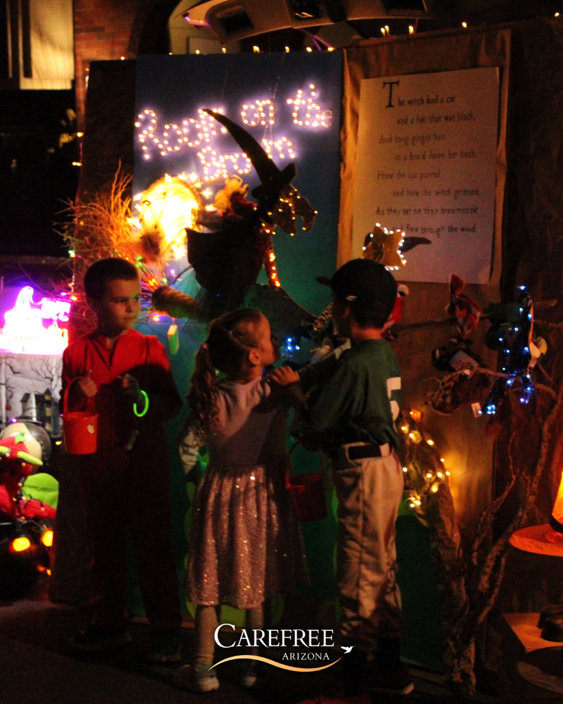 Three children in Halloween costumes gather near a decorated display with a witch figure and glowing lights at night.
