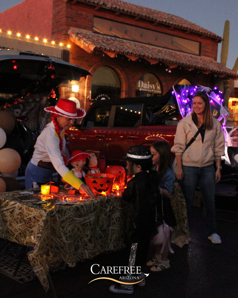 Children trick-or-treating at a Halloween-themed car trunk decorated with lights and pumpkins in front of a rustic building at dusk.