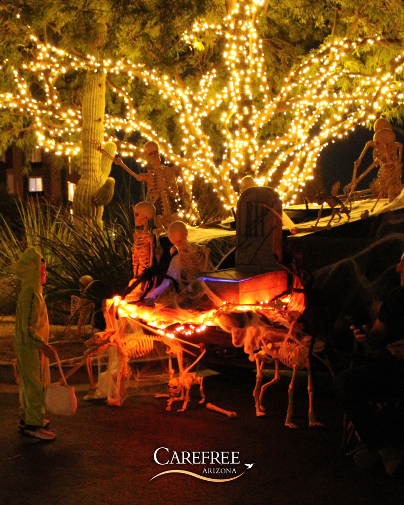 Children in costumes trick-or-treating near skeleton decorations and a tree wrapped in bright orange and white lights at night.
