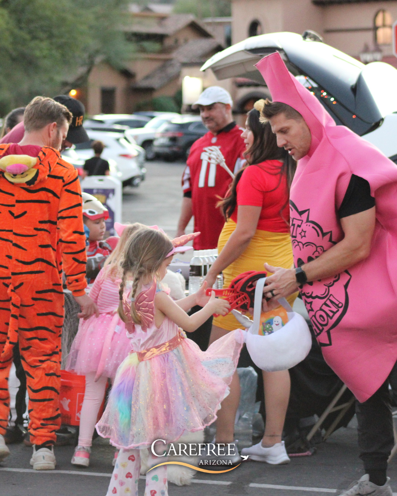 A man dressed as a whoopie cushion hands out candy to children in colorful costumes during a neighborhood Halloween event.