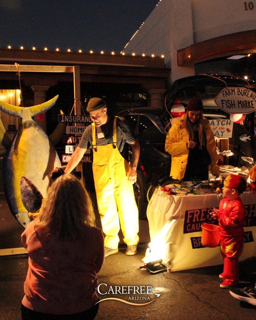 Man in yellow overalls stands beside a large fish prop at a nighttime outdoor market with a child in a red costume nearby.