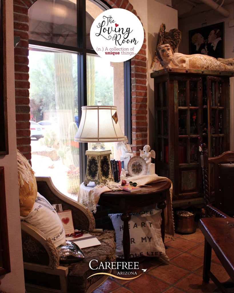 Cozy corner with vintage armchair, decorative pillows, and a lamp on a round wooden table beside a large window showing desert cacti outside.