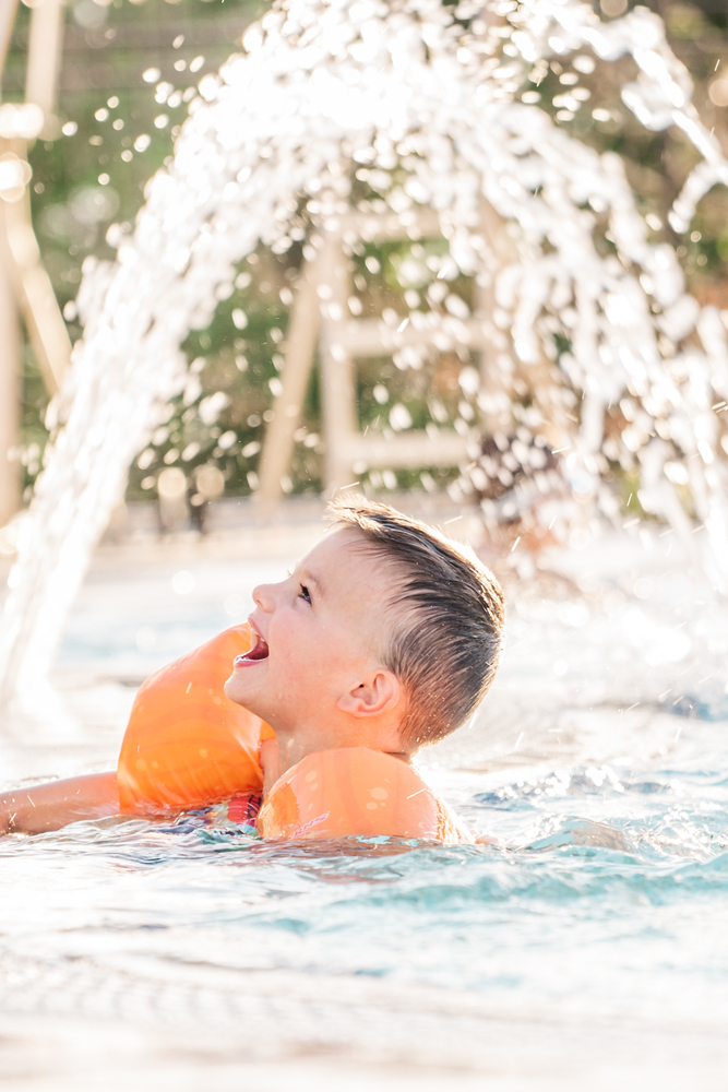 young boy in pool smiling with orang arm floaties with water splashing behind him