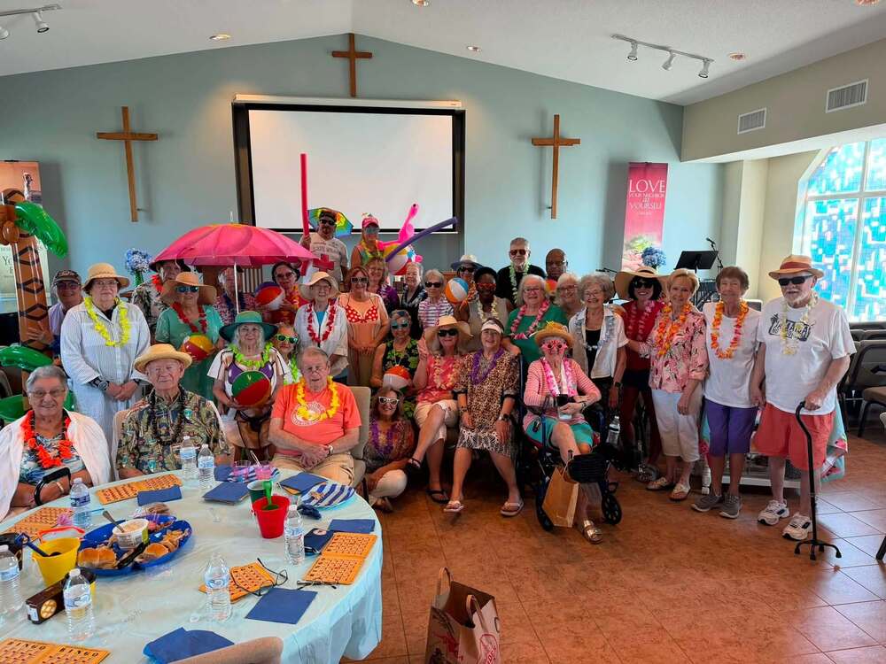 Several Senior Citizens are collected for a group photo wearing attire that gives the appearance of a luau. There is a table with food and drinks in the photo.