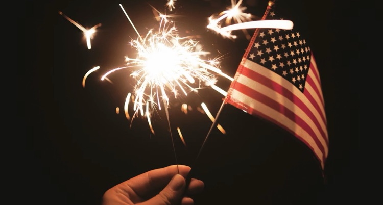 a hand holds an American flag with a sparkler to celebrate Independence Day. 