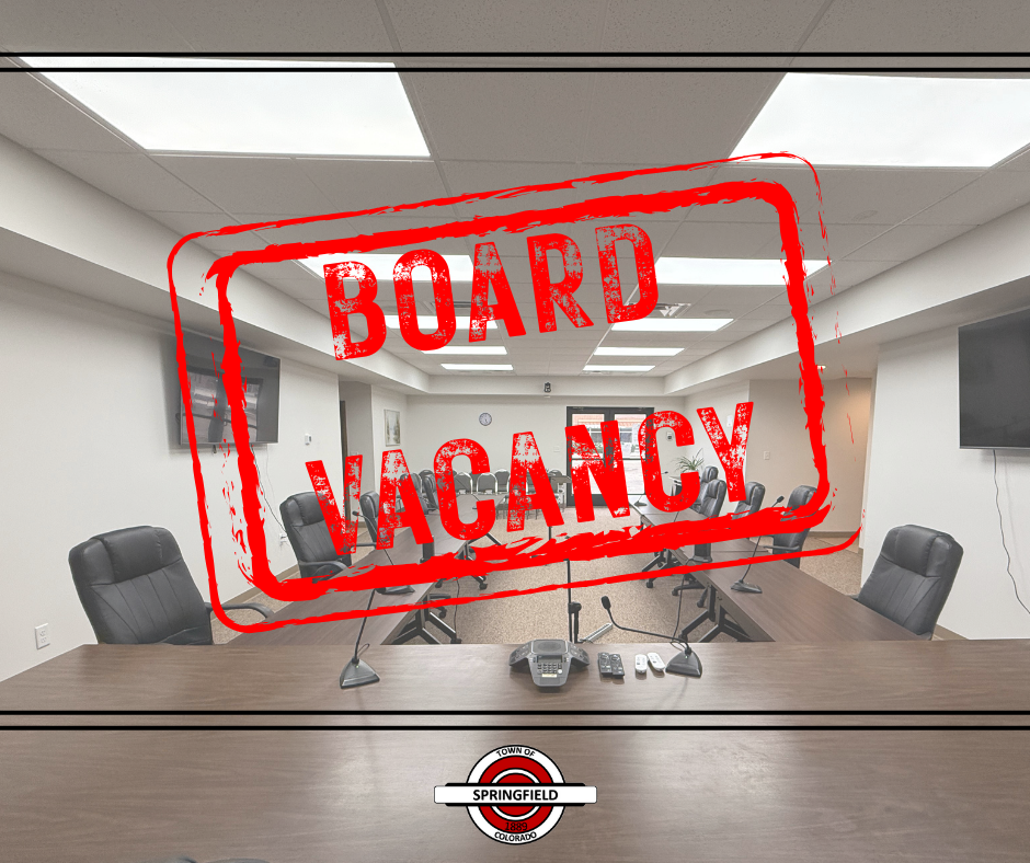 Conference room with empty chairs and microphones arranged around a large meeting table, overlaid with a bold red stamped graphic reading “BOARD VACANCY.” At the bottom is the Town of Springfield, Colorado logo.