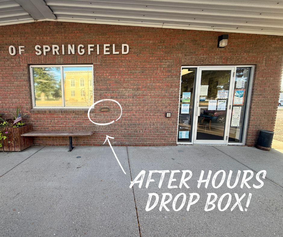 Exterior of the Springfield office building with a brick wall, window, and glass door. A white arrow points to a mail slot in the wall, labeled with large text that reads “After Hours Drop Box!”