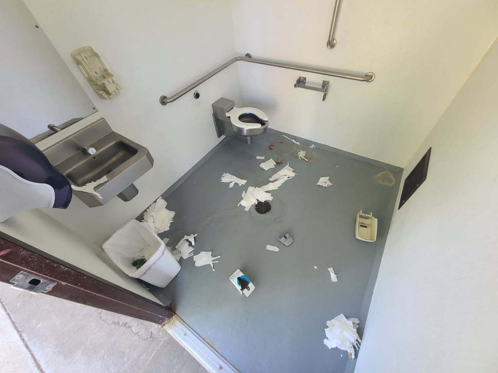 Another view of the same public restroom from a different angle. The floor is covered in scattered toilet paper, trash, and small debris. A toilet and sink are mounted along the walls with grab bars, and a trash can sits nearby. Despite available disposal options, waste is spread across the floor, creating a messy and unsanitary scene.