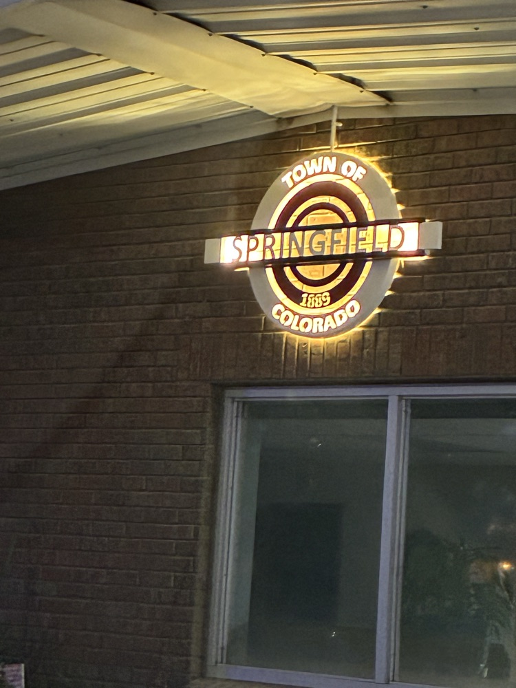 Exterior of a brick building at night with a “Town of Springfield, Colorado” sign illuminated above the entrance.