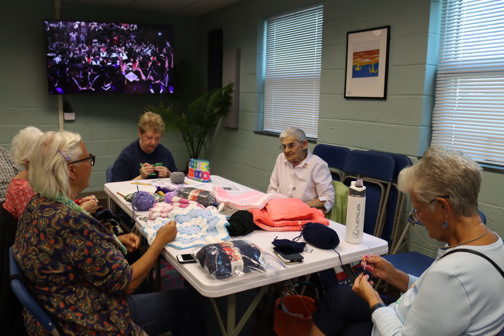 Group of women sitting around a table knitting while a tv plays in the background of the lounge