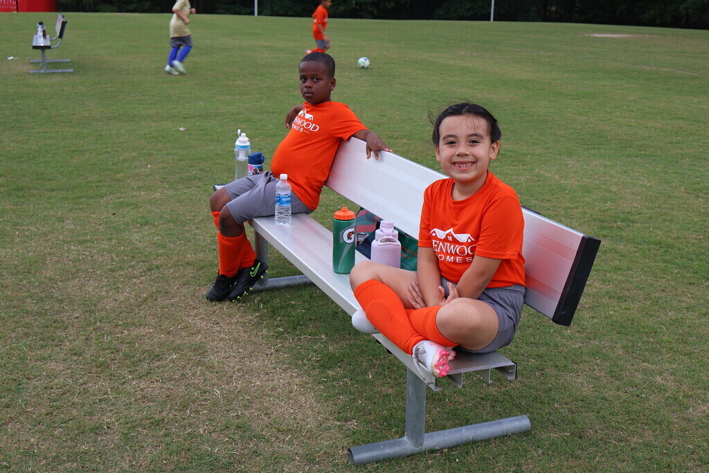 Two children in soccer uniforms sitting on the bench with water bottles