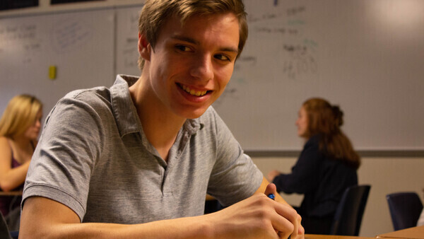 young male teen smiling at camera while sitting in class with class mates in the background