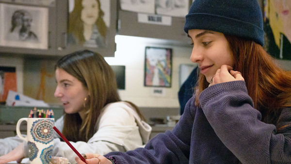 two female students in art room with pencils in hand while sitting beside each other at table