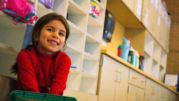 image of little girl smiling at camera in classroom wearing red shirt.