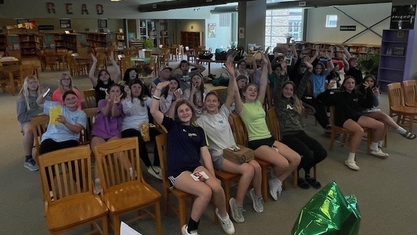 image of several students in library raising hands and smiling at camera.