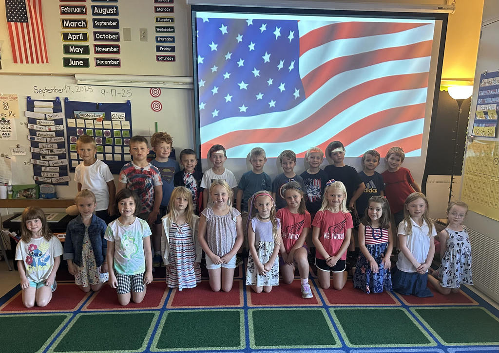 students knealing on the classroom floor in front of  a screen with an American flag image on it